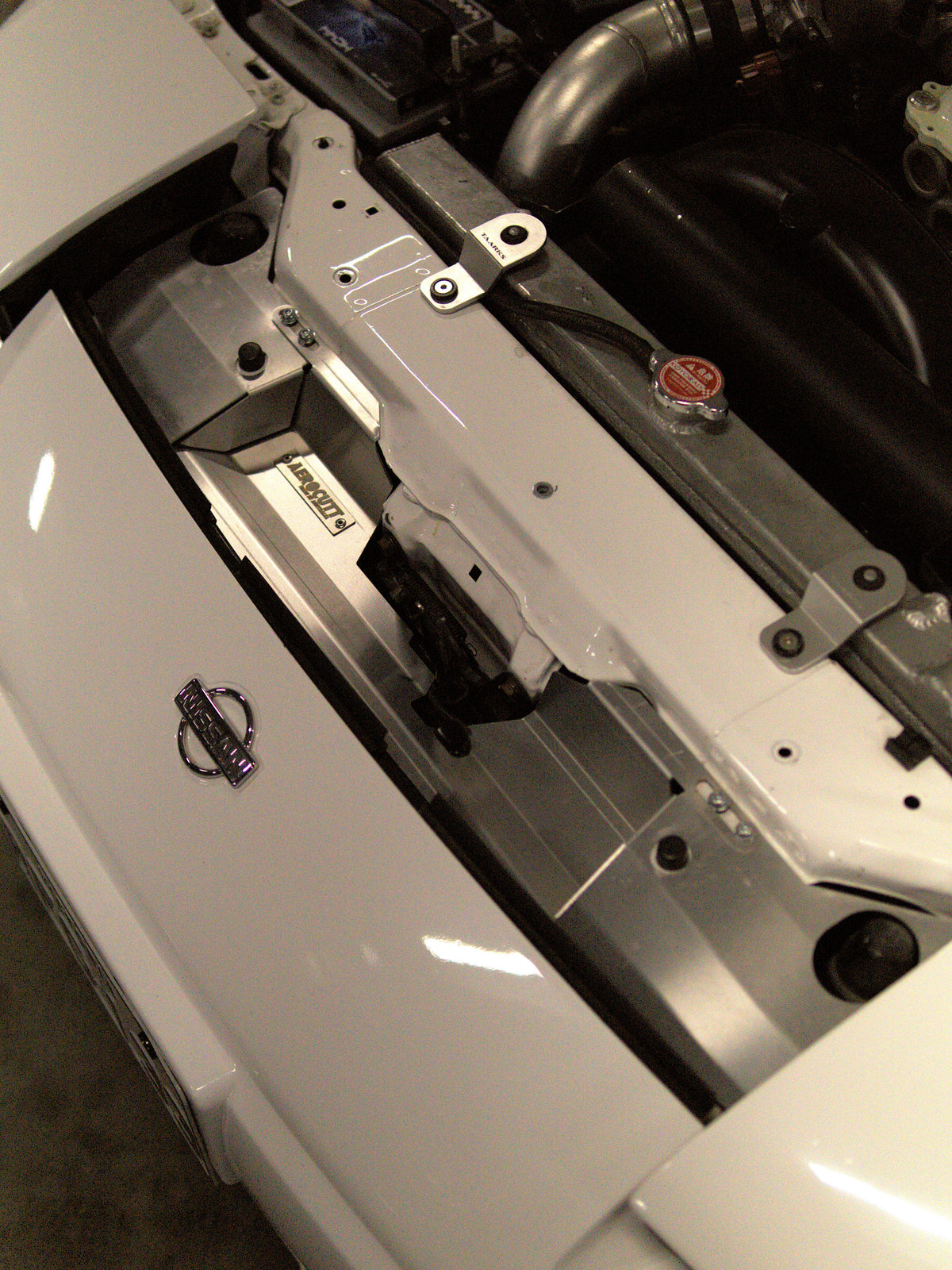 Close-up of a white car hood opening, showing the engine bay with a silver metal 180sx aerocutt cooling panel set installed on the inner fender area. A small black and purple Nissan logo is visible on the white hood.