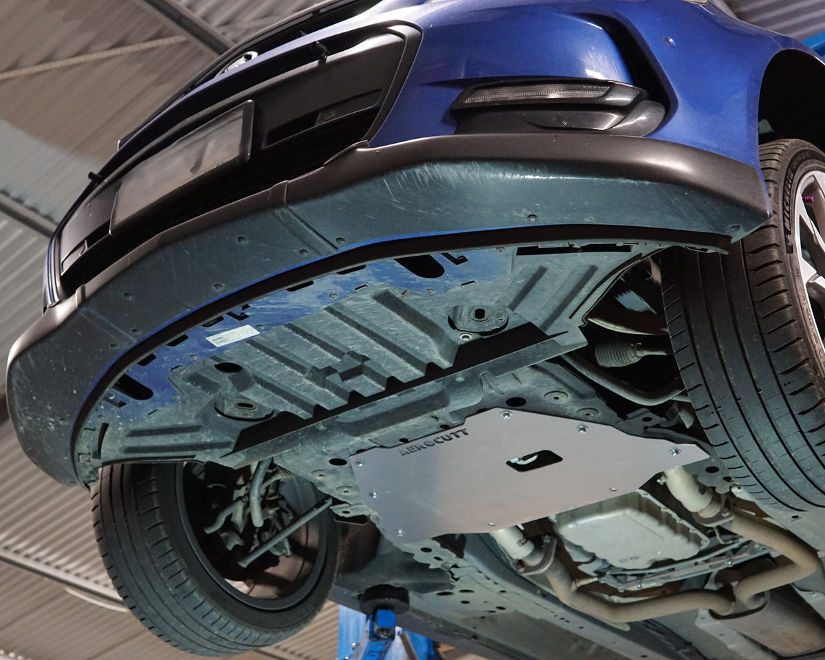 A silver metal underbody shield installed on the chassis of a blue car, photographed from underneath as the vehicle is raised on a lift in a garage.