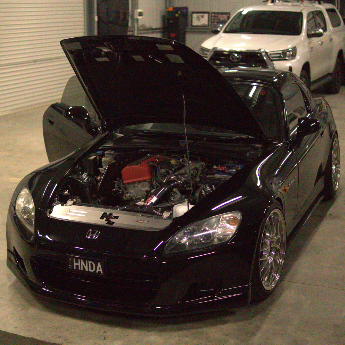 A glossy black Honda S2000 with its hood open, showcasing the engine bay with an Aerocutt cooling panel fitted