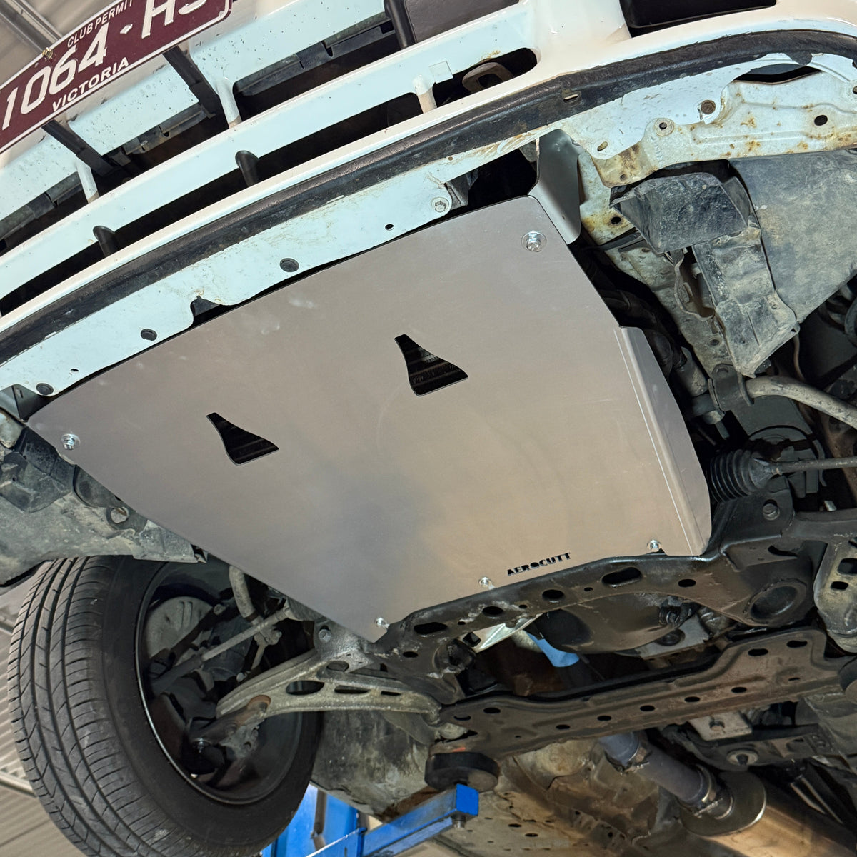 Metal skid plate on a vehicle chassis in a workshop setting, mazda rx7 fc underneath engine bay 